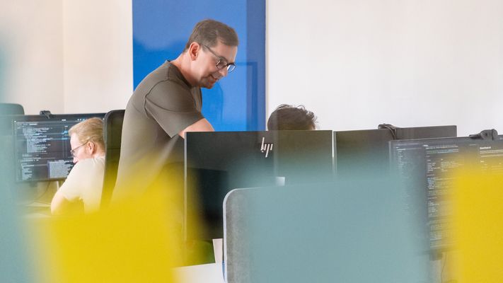 Büro mit Arbeitsplätzen, ein Kollege beugt sich über einen Schreibtisch.