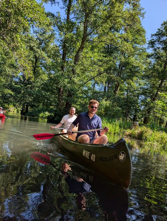 Zwei Kollegen paddeln im Kanu durch einen von Bäumen gesäumten Wasserlauf.