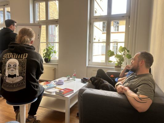 Runde im Büro - zwei stehen am Flipchart, zwei sitzen auf dem Sofa, ein Hund liegt dazwischen, auf dem Tisch Fachbücher.