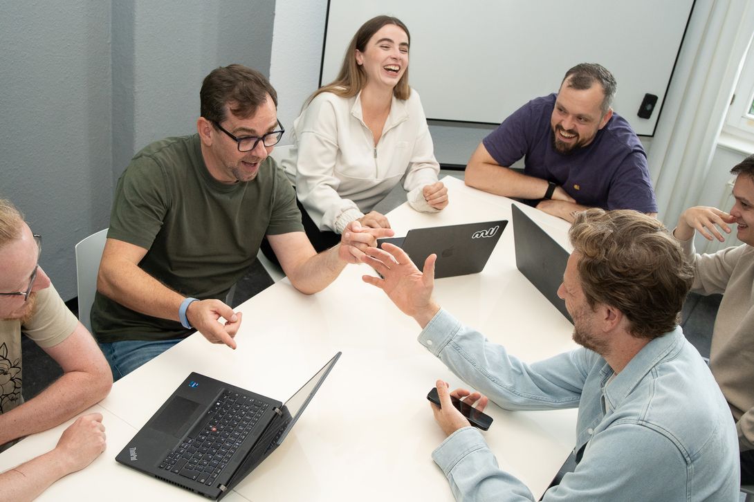 Sieben mustworker:innen sitzen um einen Besprechungstisch mit aufgeklappten Laptops und lachen.