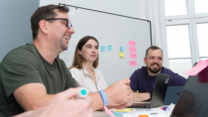 Drei aus dem Team am Besprechungstisch, einer lacht, dahinter ein Whiteboard mit Haftnotizen.