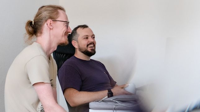 Zwei Kollegen lachen gemeinsam am Schreibtisch, einer steht, einer sitzt im Bürostuhl.